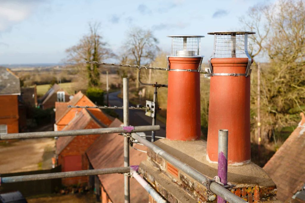 Two red brick chimney pots with mesh covers on a rooftop, surrounded by scaffolding. In the background, there are trees, buildings, and a country road under a blue sky.