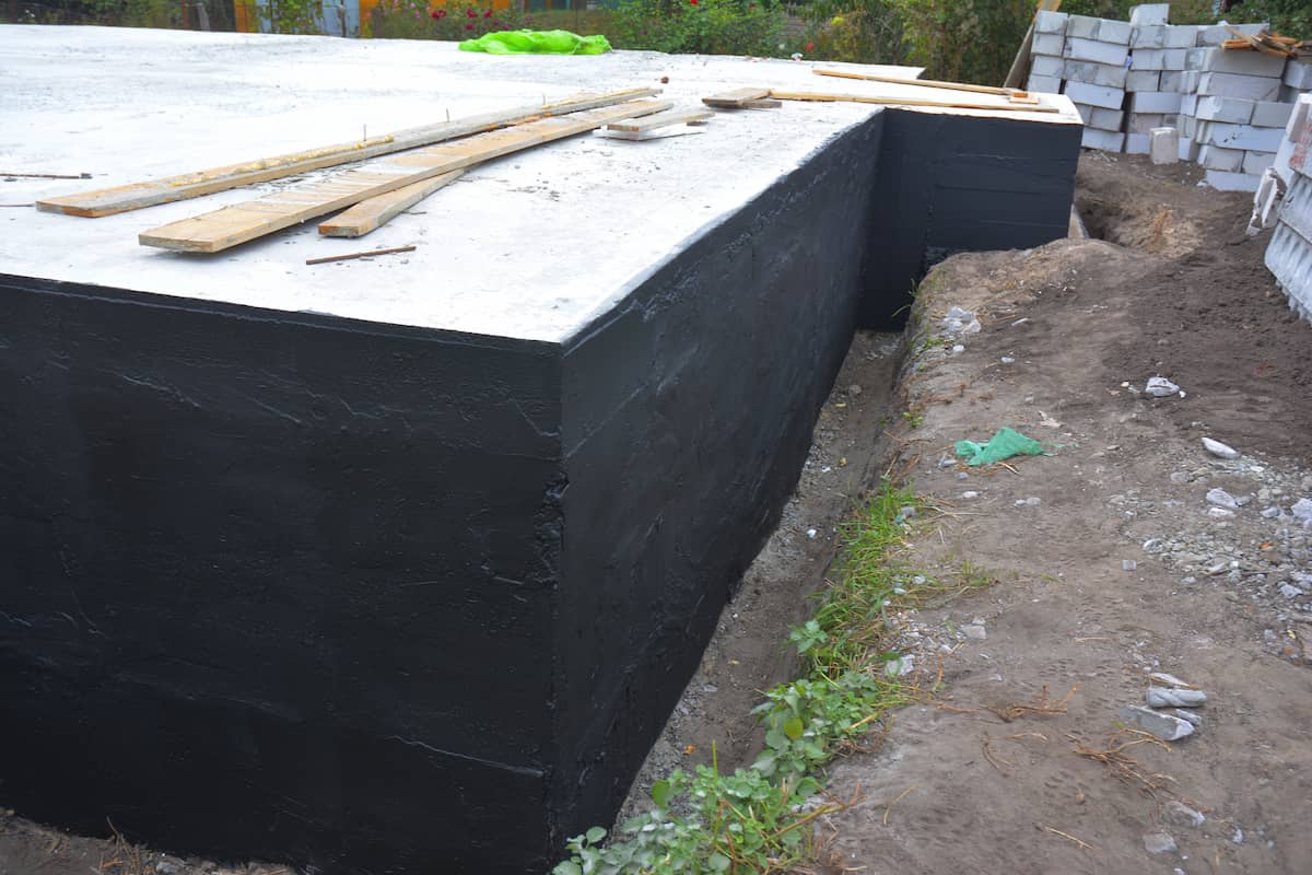 Concrete slab foundation with black waterproofing on the side, surrounded by soil and construction materials, wooden planks lying on top of the slab.