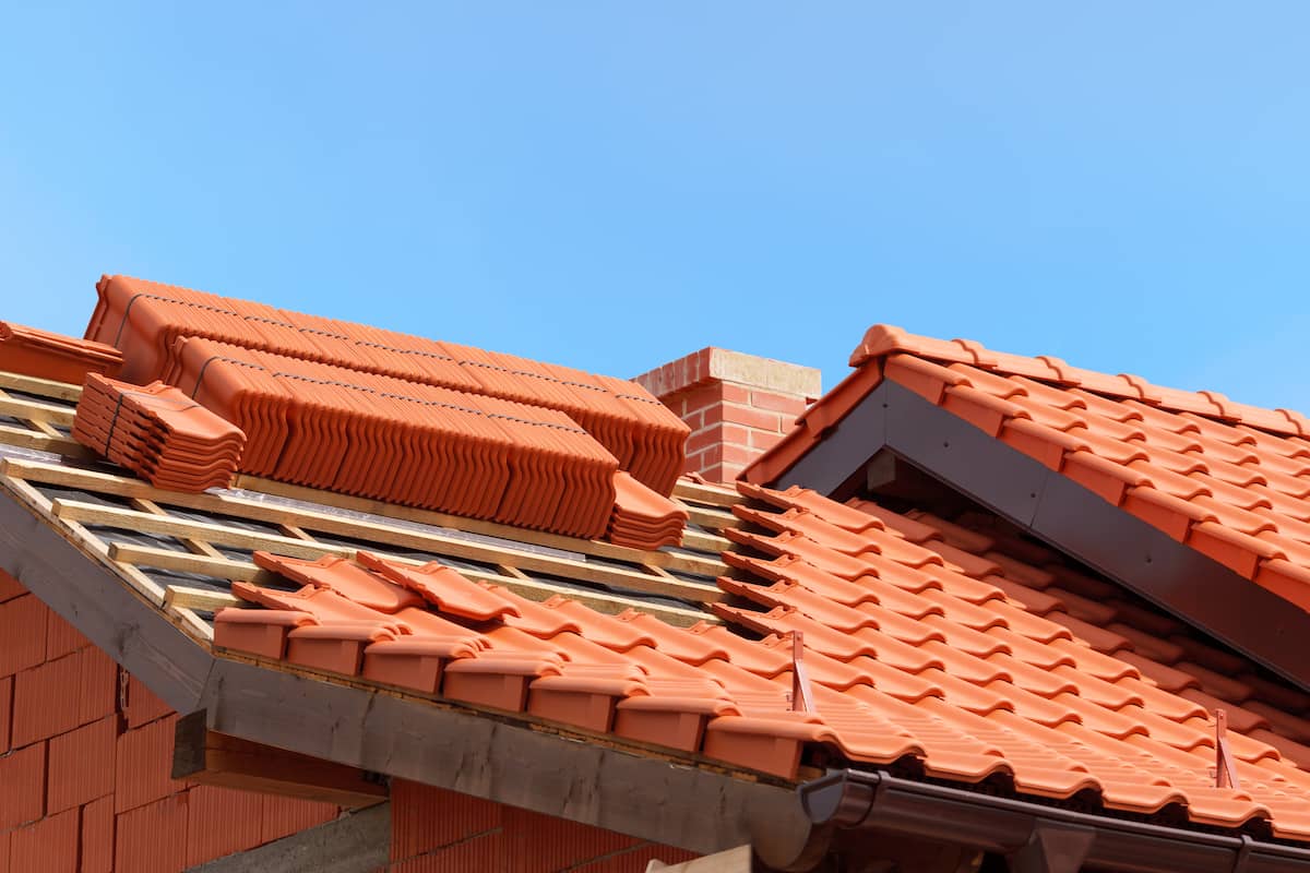 A house roof under construction with red clay tiles, some already installed and others stacked nearby. A brick chimney is visible, and the sky is clear and blue.