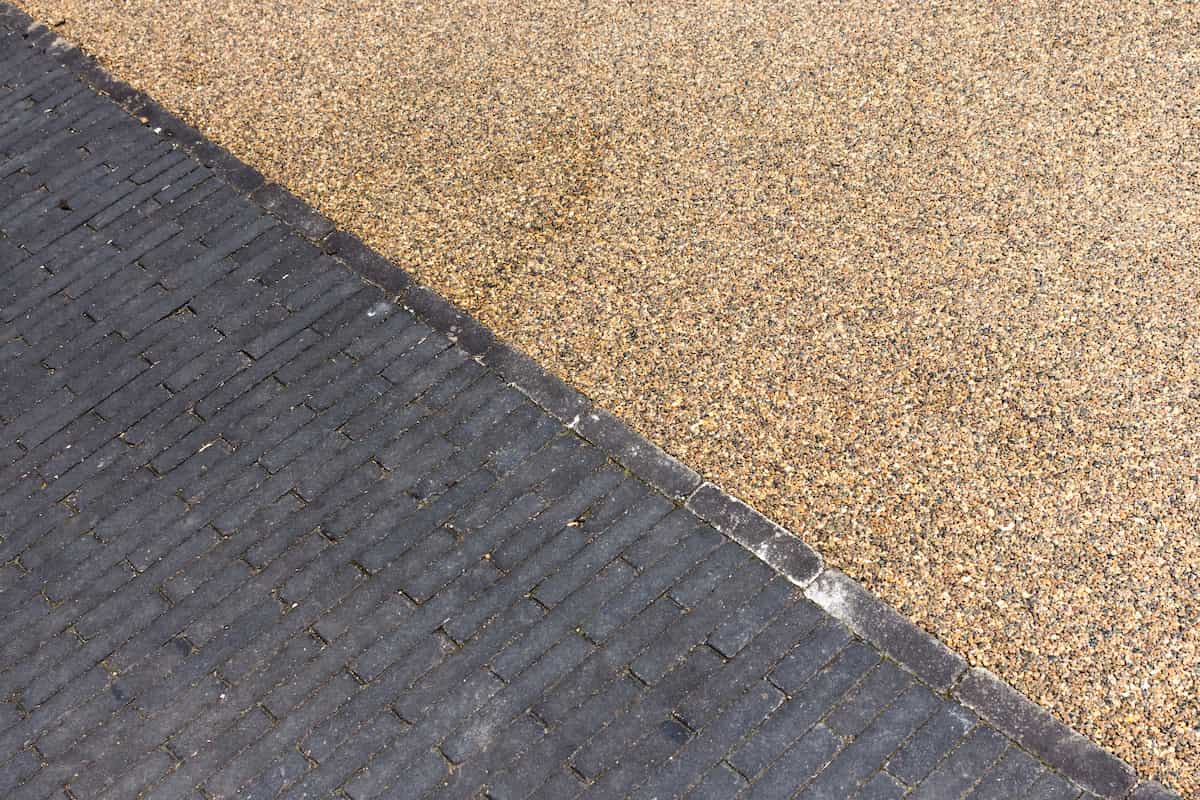 A diagonal line separates a dark gray brick pavement on the left from a light brown gravel surface on the right.
