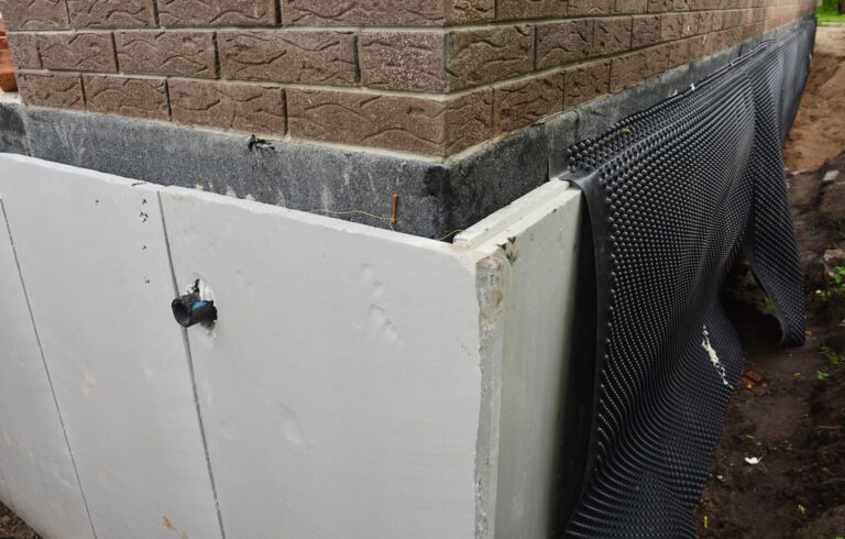 A close-up of a building foundation under construction, showing white insulation panels, black waterproofing membrane, and brick exterior walls above. Soil is visible beside the foundation.