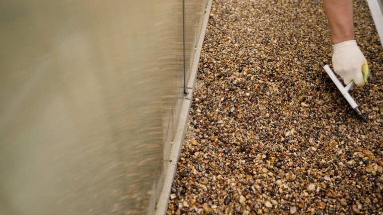 A person wearing a white glove uses a trowel to smooth small pebbles along the edge of a wall.