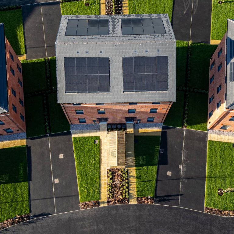 Aerial view of a brick building with solar panels on the roof, surrounded by green lawns, pathways, and curved black asphalt roads.