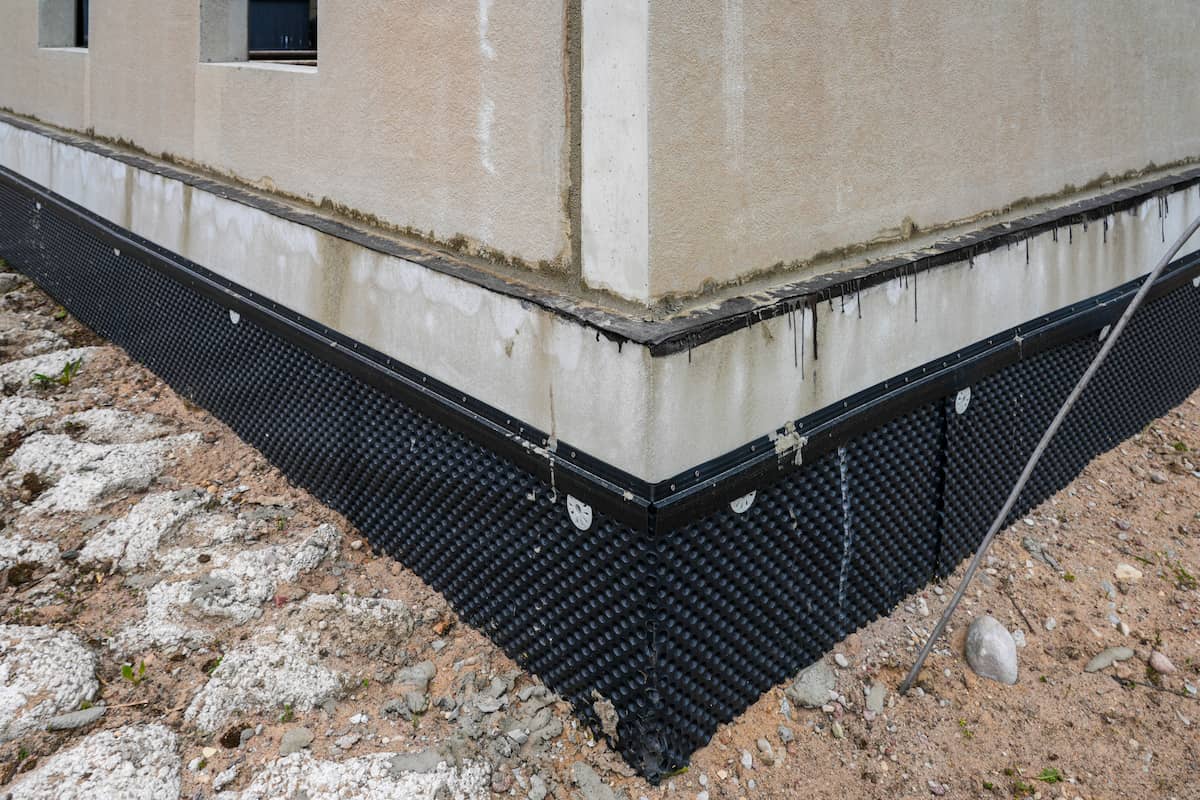 The image shows the foundation of a building with waterproofing membrane installed. The wall has stains and the ground around the foundation is rough, with exposed soil and rocks.