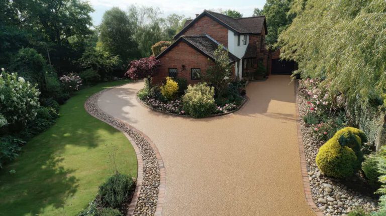 A brick house with a pitched roof is surrounded by gardens, featuring winding stone-edged paths, trimmed bushes, green grass, colorful flowers, and tall trees.