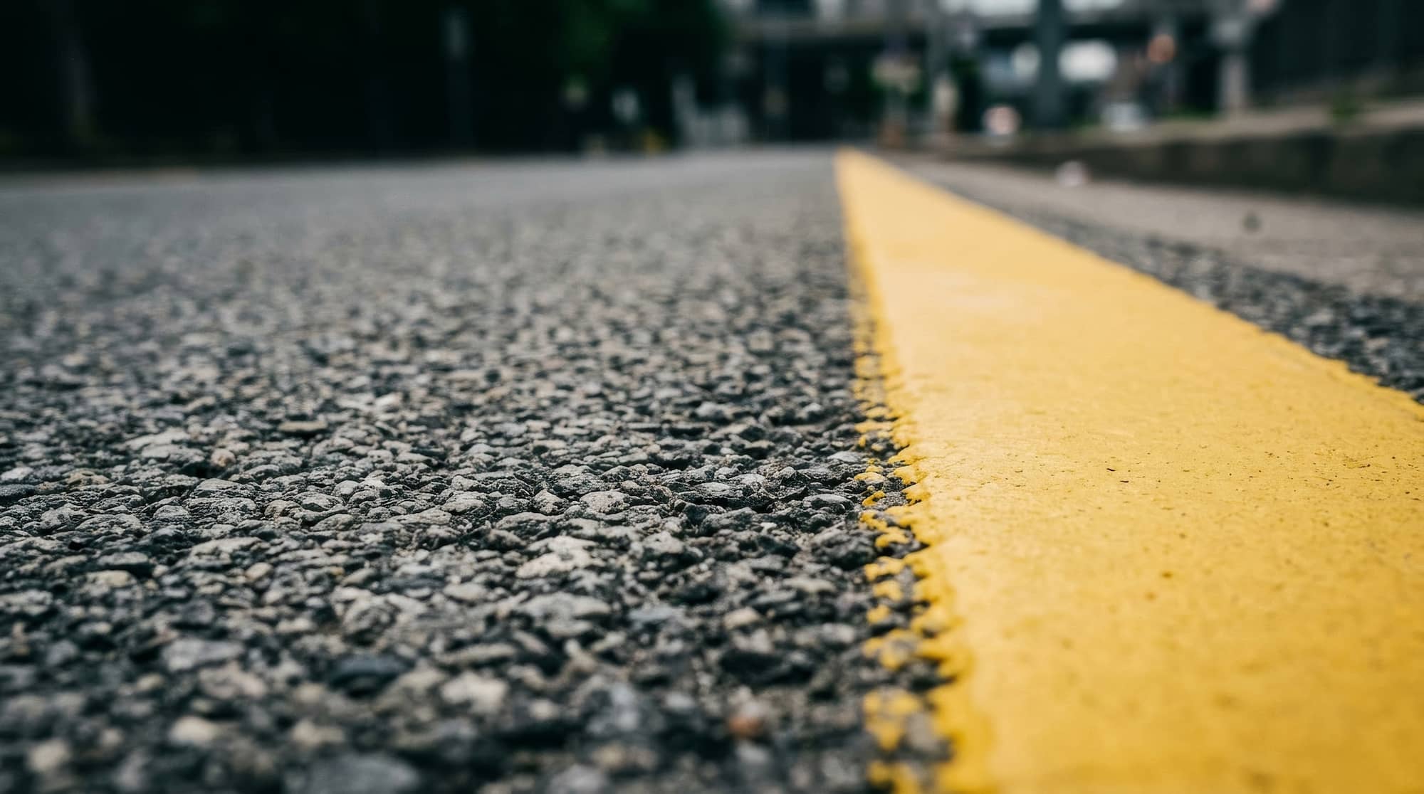 Close-up view of a rough asphalt road with a single, bright yellow line running through the middle. The focus is low to the ground, with the background blurred and out of focus.