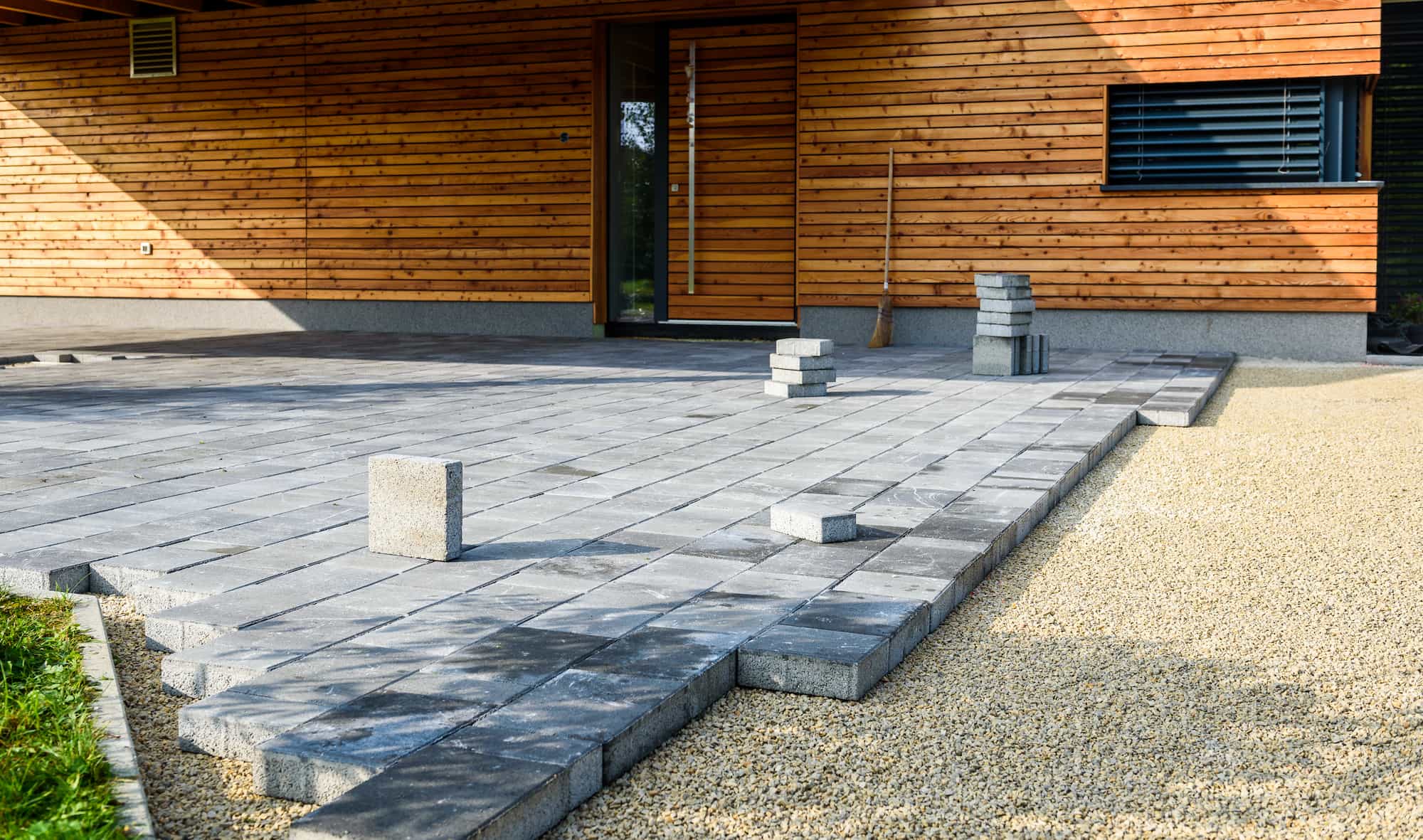 A patio made of gray concrete pavers is partially constructed next to a wooden building. Several stacks and loose pavers are visible, with gravel surrounding the patio area.
