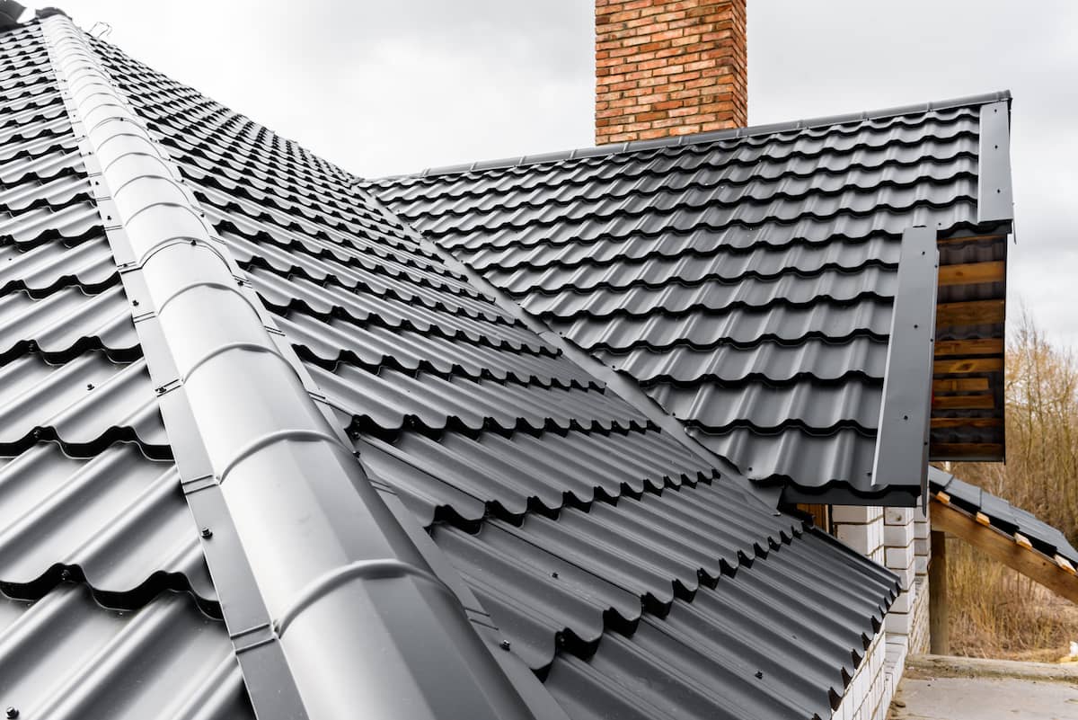 Black metal roofing with a wavy pattern on a house, brick chimney, and cloudy sky in the background. Some trees and partial building structure are visible.