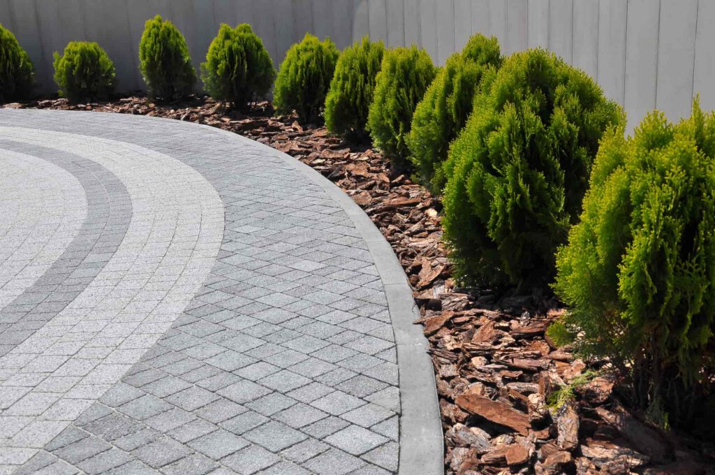 Curved, gray stone-paved path bordered by small, neatly trimmed green shrubs and brown wood chips, next to a white fence.