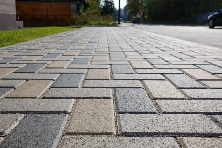 Close-up view of a paved sidewalk with a brick pattern, taken from a low angle. The walkway extends into the distance, with grass on one side and a blurred figure far ahead.