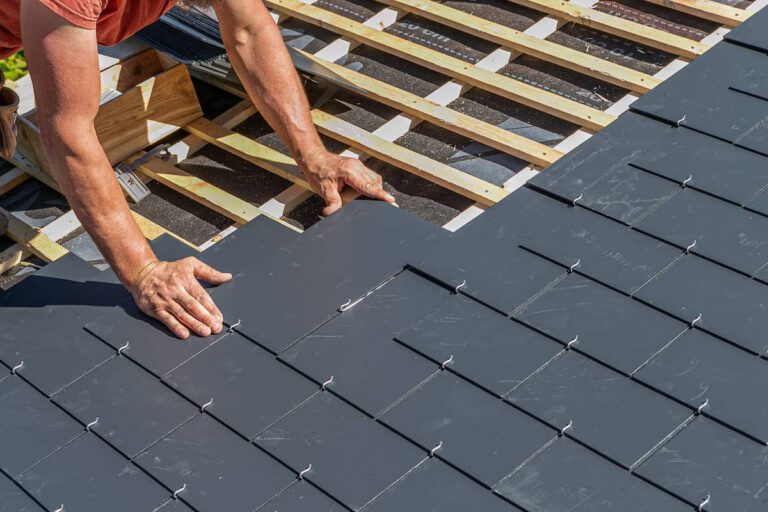 A person installs rectangular gray slate tiles on a roof, arranging them in rows over a wooden framework under bright daylight.