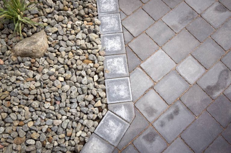 Gray patio pavers meet a border of square stones, separating the paved area from a section of small gray rocks with a few green plants and a large rock visible.