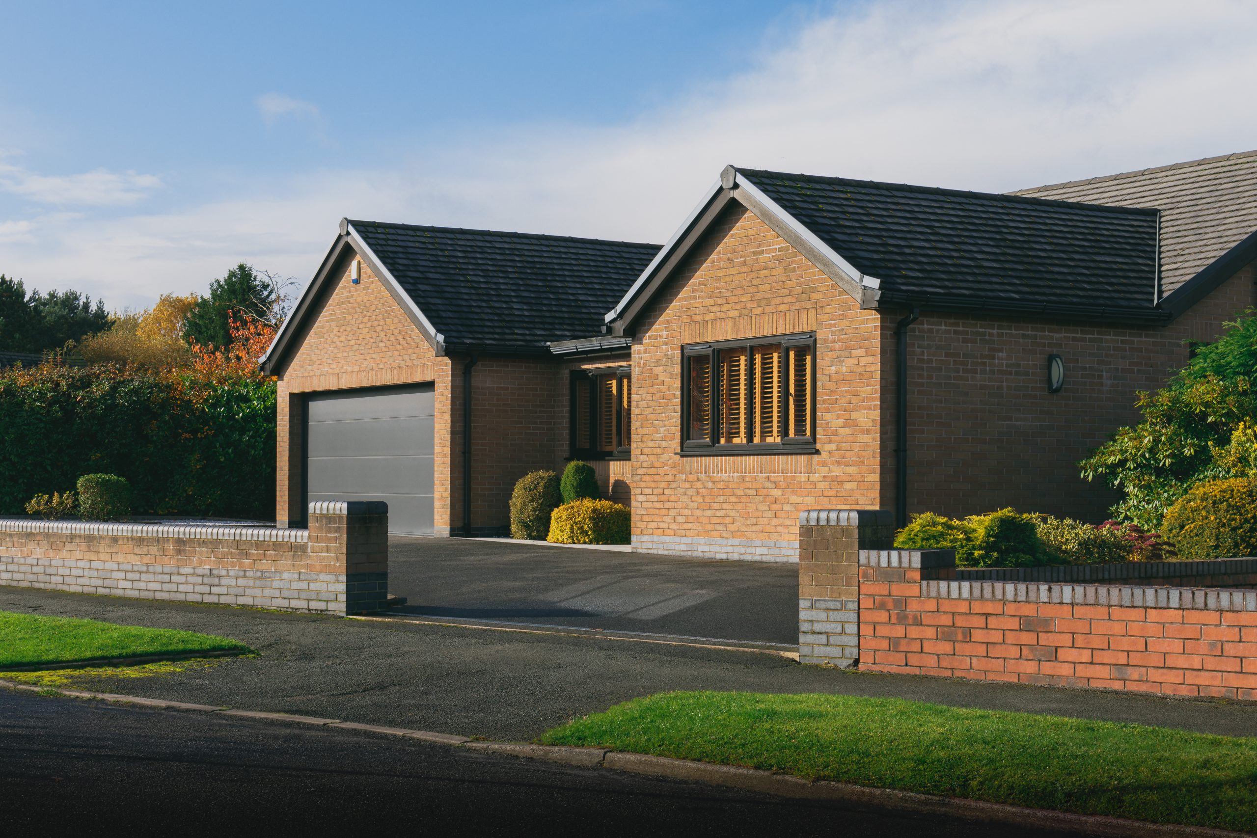 A modern, single-story brick house with a dark gray double garage, neatly trimmed bushes, a low brick wall, and a driveway, all under a blue sky with some clouds.
