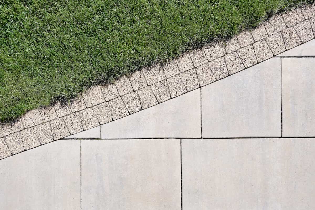 Diagonal view of green grass bordered by small square stones, next to large rectangular concrete pavement tiles, creating a geometric, contrasting pattern.
