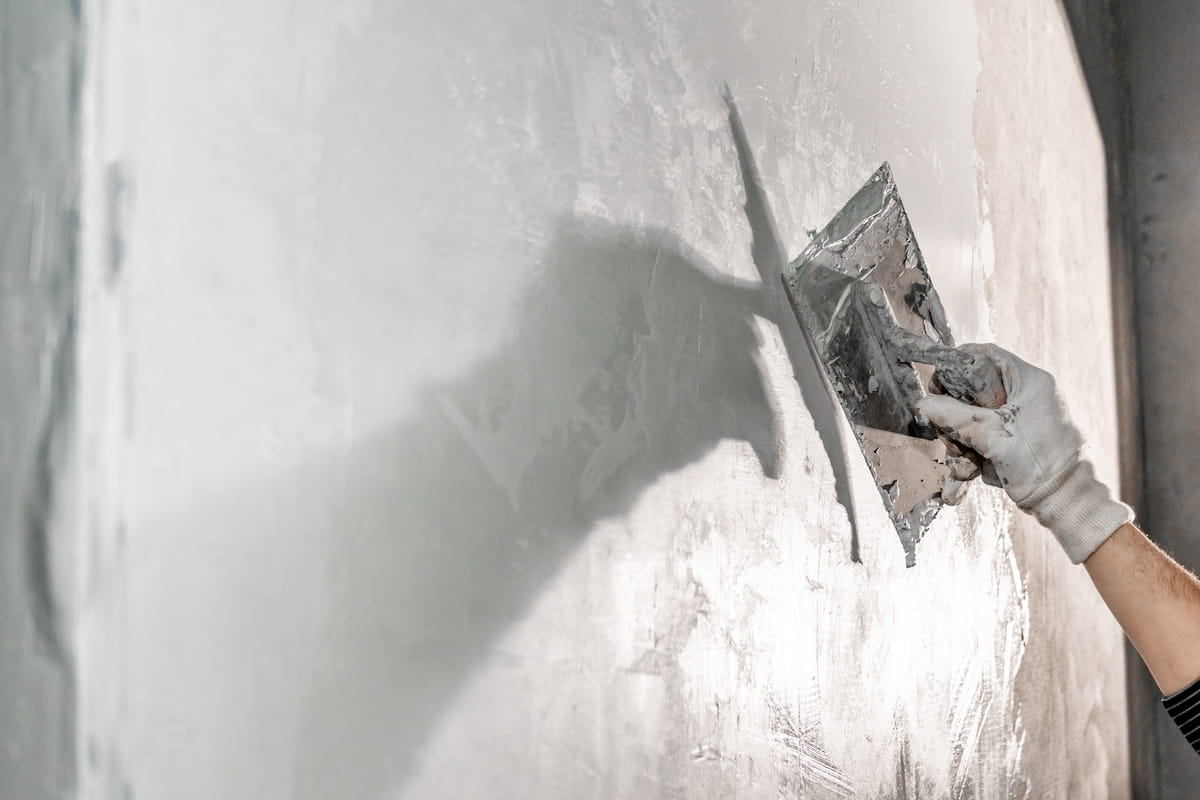A gloved hand smooths plaster on a wall with a trowel, casting a shadow.