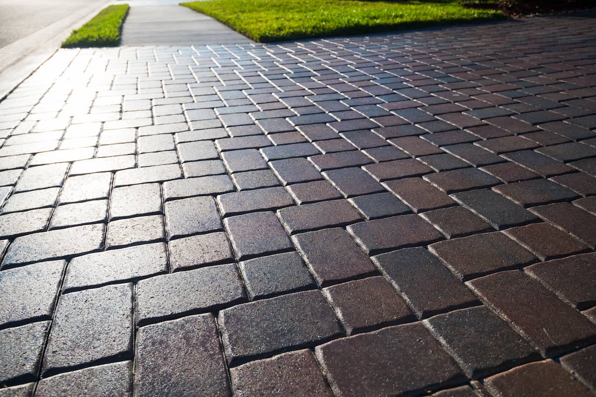 Close-up view of a sunlit brick paver sidewalk next to a green grassy area, with the edge of a street visible in the background.