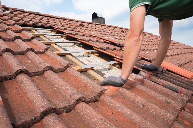 A person in socks repairs a tiled roof, with several red tiles removed to reveal wooden beams and insulation underneath. The person is kneeling, working with their hands on the roof.