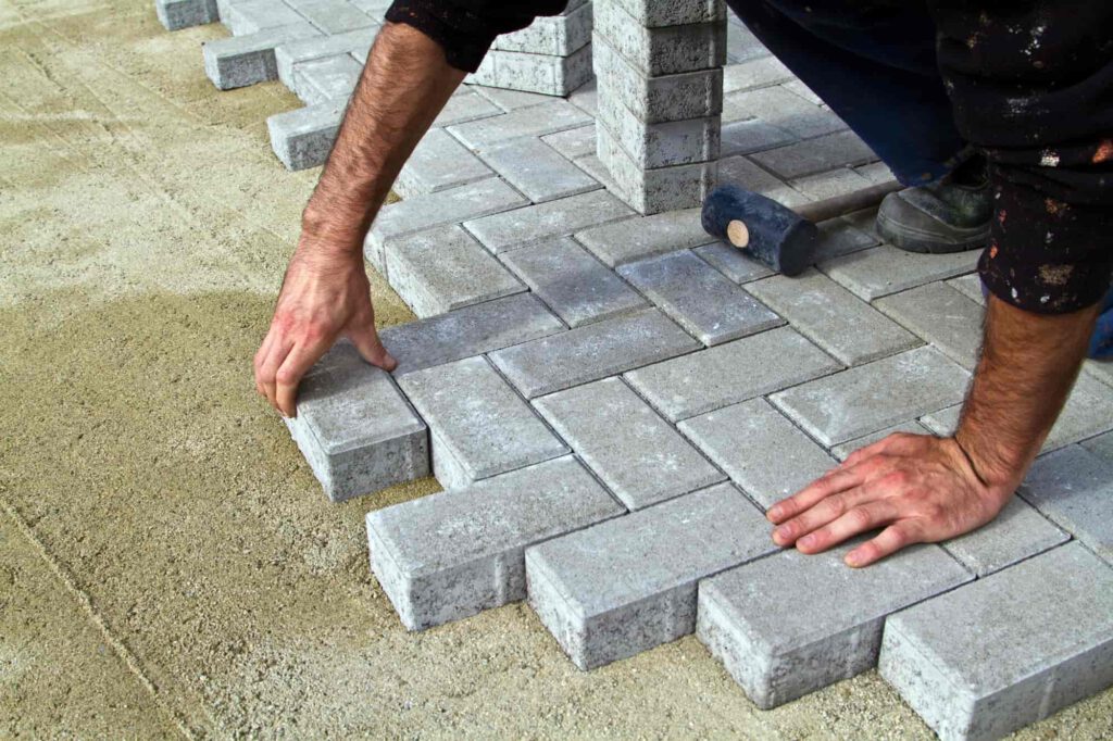 A person is laying rectangular gray paving stones in a herringbone pattern on a sandy surface, using their hands to position the bricks. A rubber mallet is nearby.