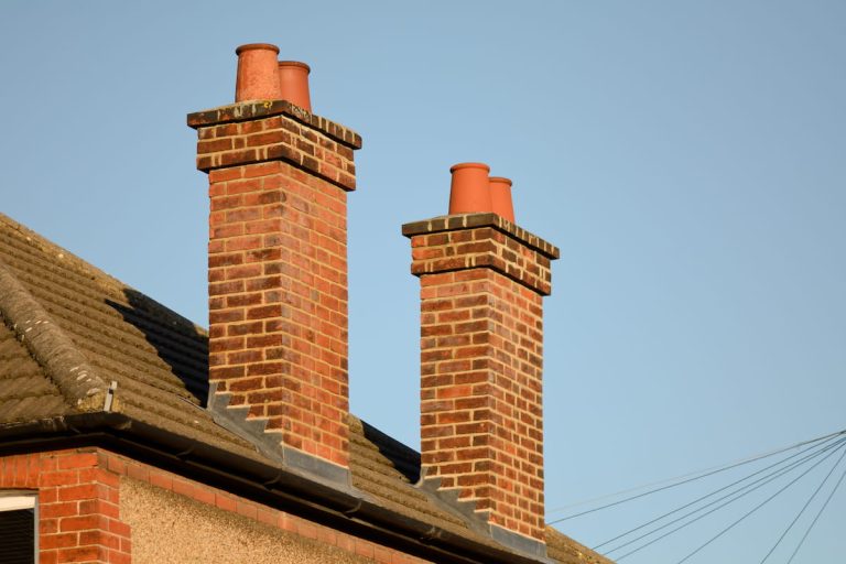 Two red-brick chimneys with clay chimney pots on a house roof, set against a clear blue sky. Roof tiles and part of the building are visible in the foreground.
