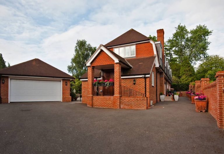 Large brick house with dark roof, a separate double garage, paved driveway, potted plants along the walkway, and a brick wall bordering the property. Trees and greenery surround the home, and the sky is partly cloudy.