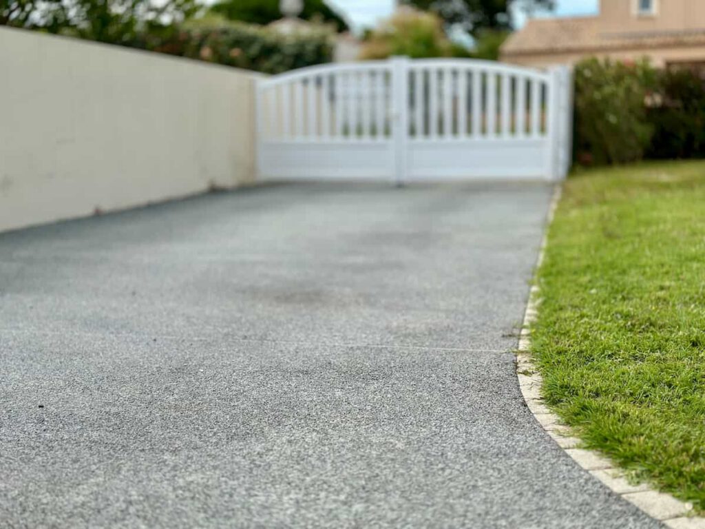 A close-up view of a paved driveway curves alongside green grass, leading to a closed white gate and a cream-colored wall in a residential area.